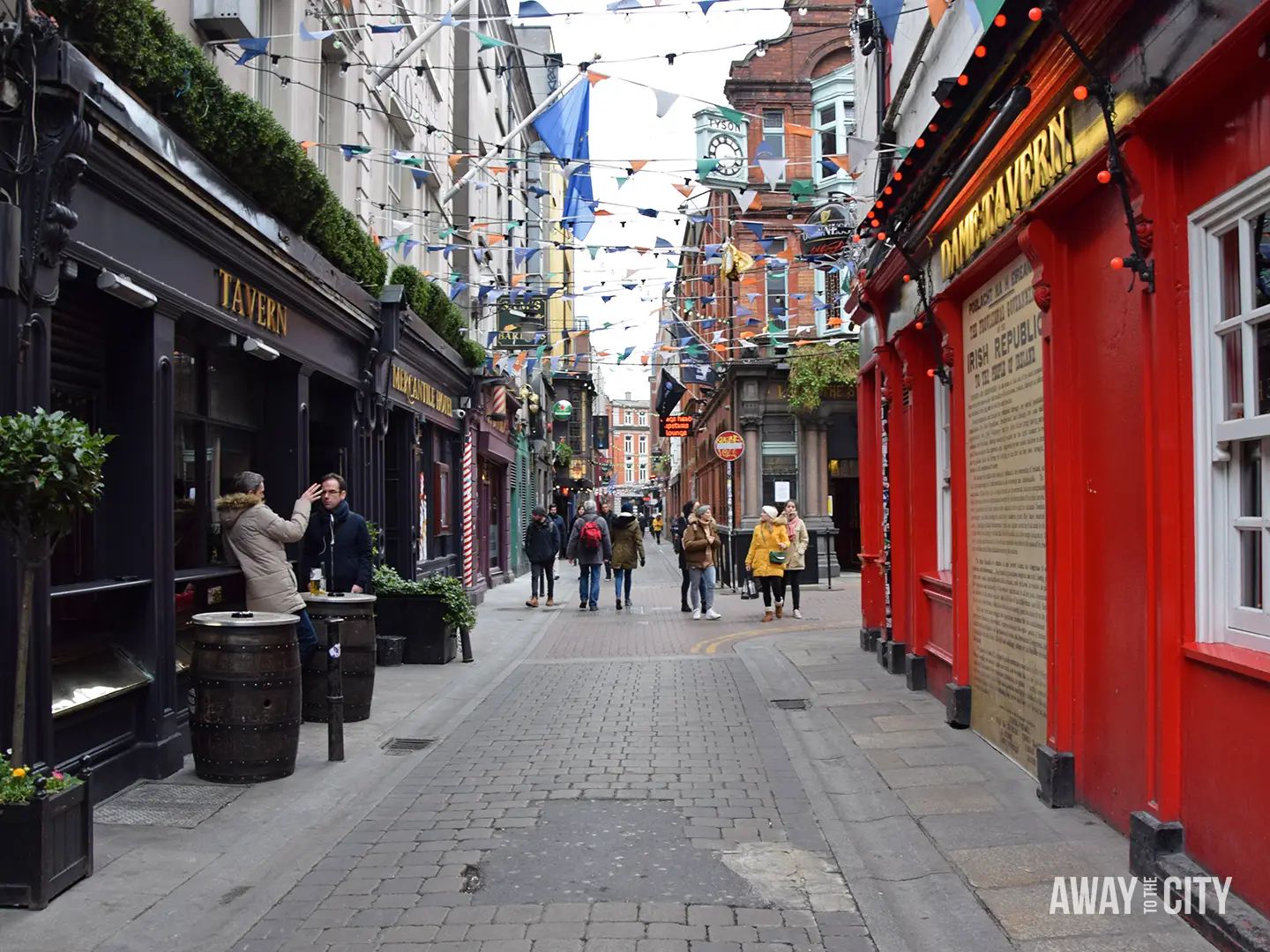 Narrow Dublin lane in Temple Bar with pubs, colourful bunting overhead, pedestrians walking between dark tavern fronts and bright red façades.