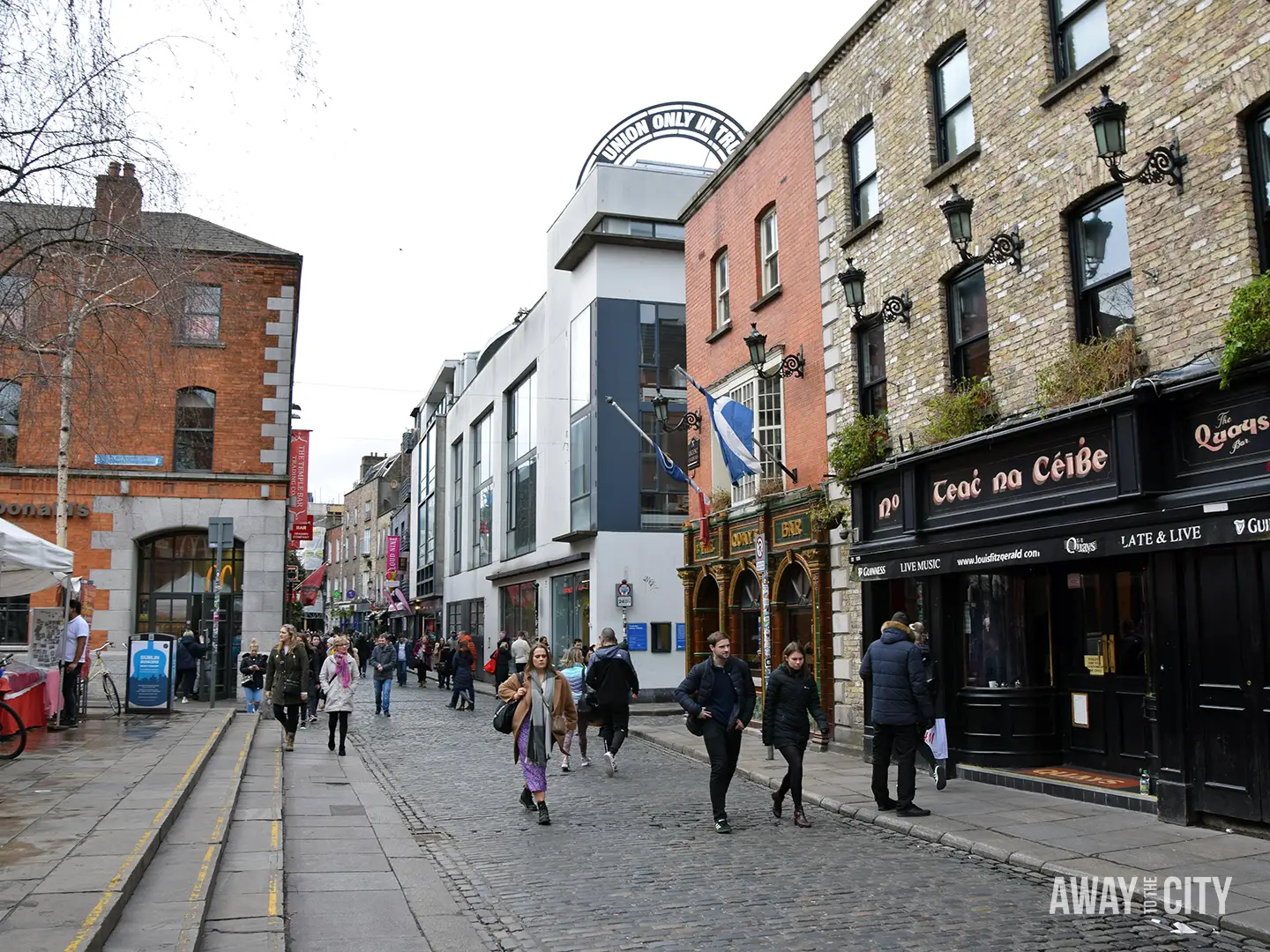 Busy cobbled street in Dublin with pedestrians, traditional pubs and shopfronts, featuring signs for Teach na Ceibe and surrounding brick buildings.