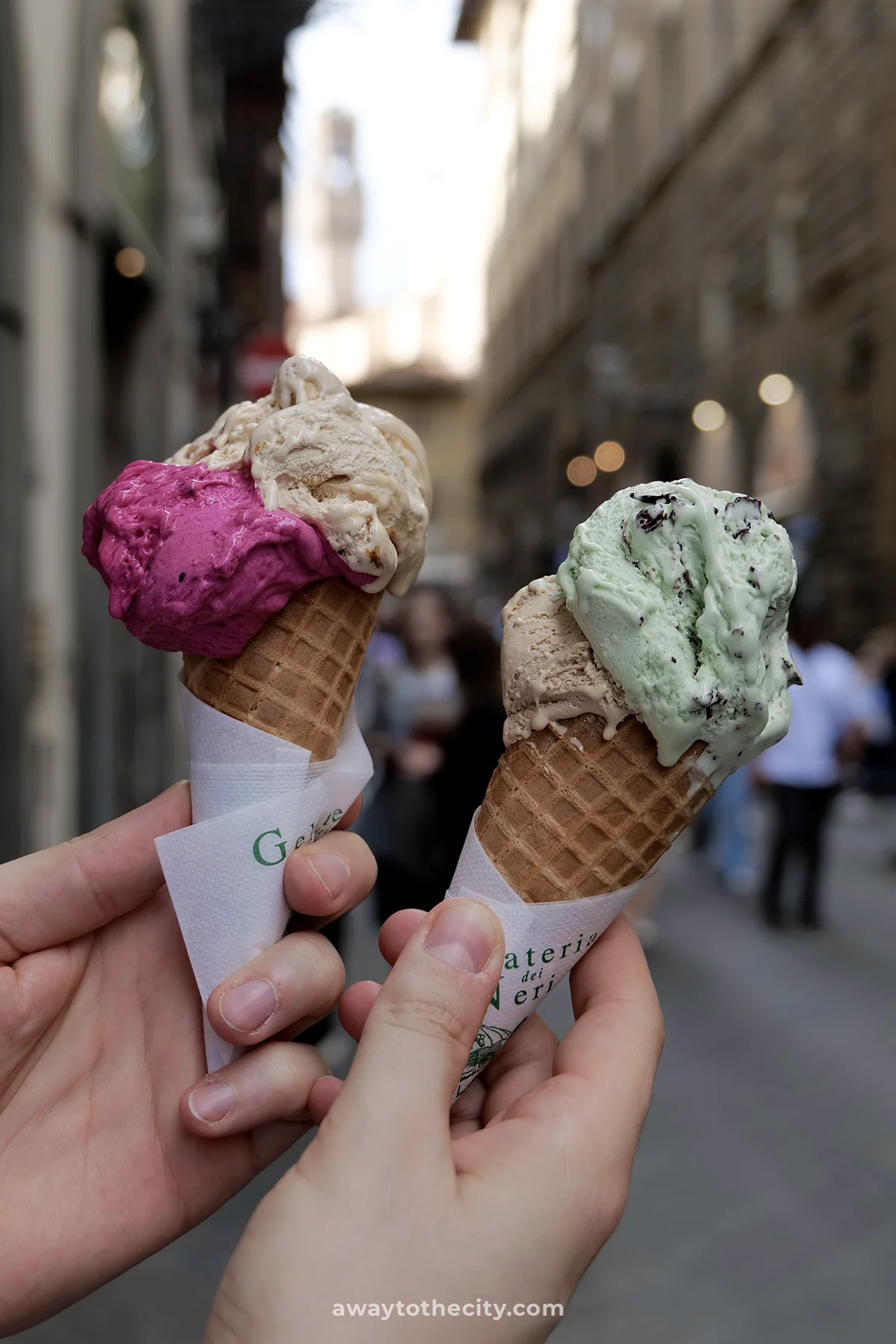 Two ice cream cones, one with pink and the other with mint chocolate chip gelato, are held up against a blurry Florence street backdrop.