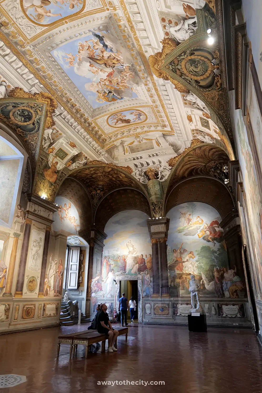 A visitor sits on a bench in the opulent Treasury of the Grand Dukes at Pitti Palace, surrounded by intricately painted walls and ceilings.