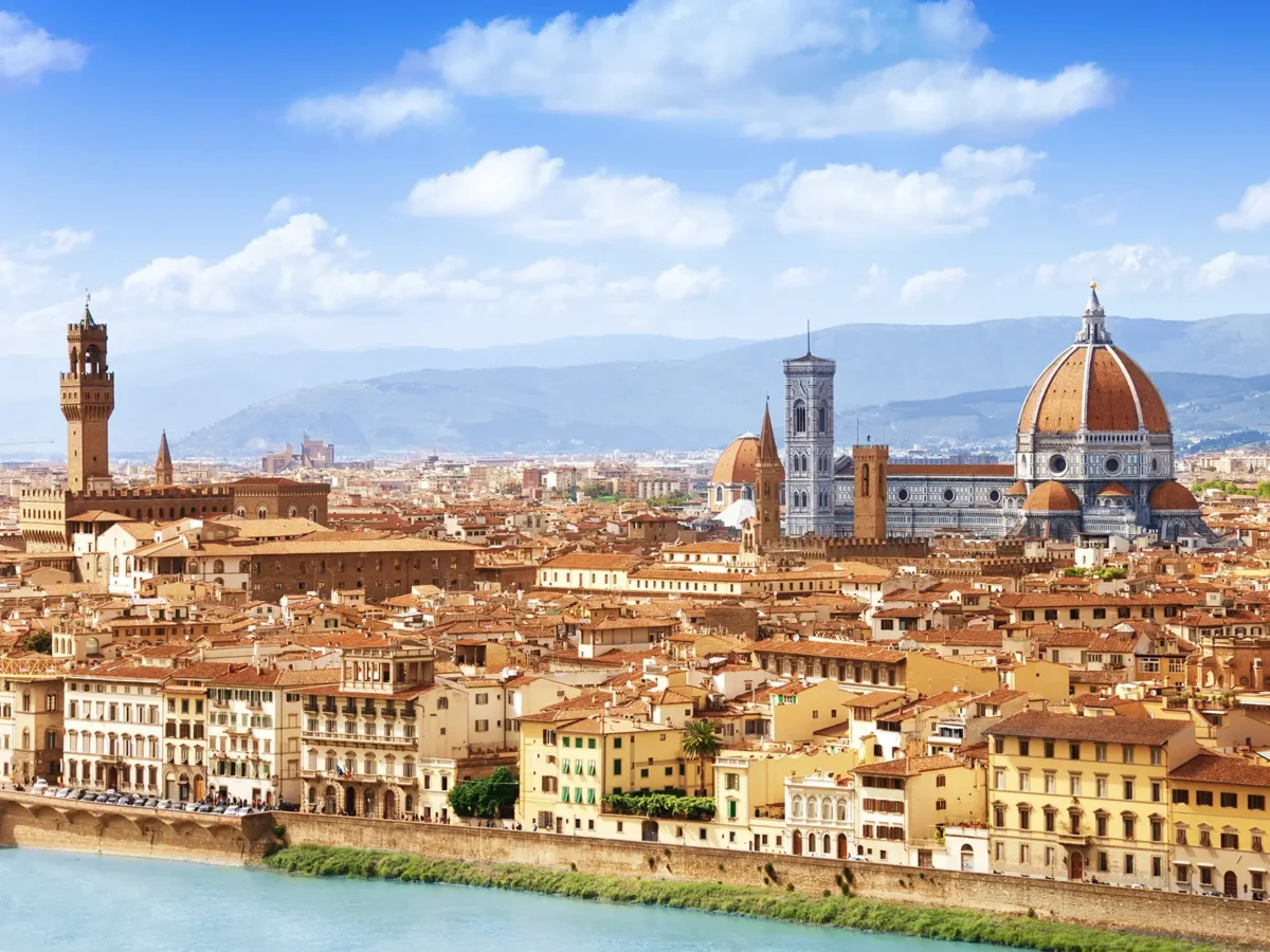 Florence's cityscape is dominated by the Duomo, a large cathedral with a distinctive red-tiled dome, and other historic buildings, all under a bright blue sky.