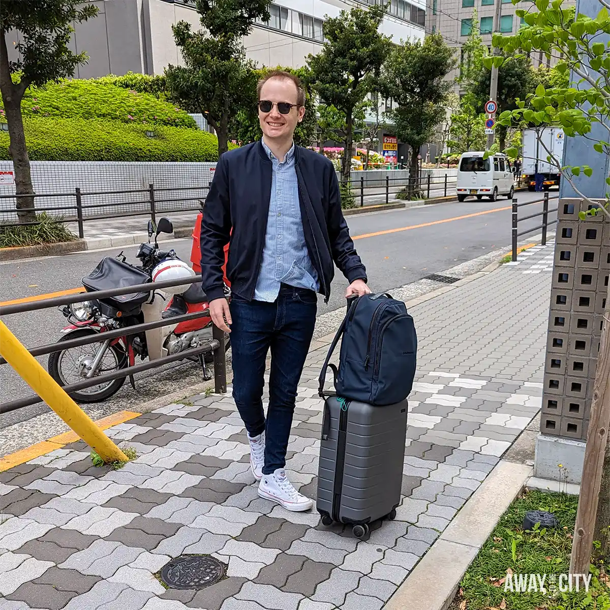 Person walking along a pedestrian pavement in Osaka with a wheeled suitcase and backpack, beside a quiet road lined with greenery.