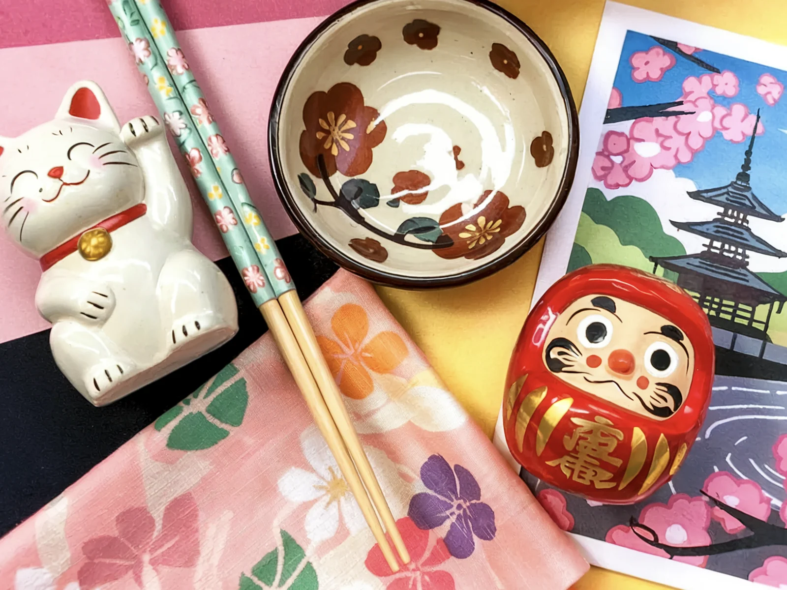 A collection of small Japanese items, including a Maneki Neko, chopsticks, a bowl, a Daruma doll, and a cloth, are arranged together as potential souvenirs.