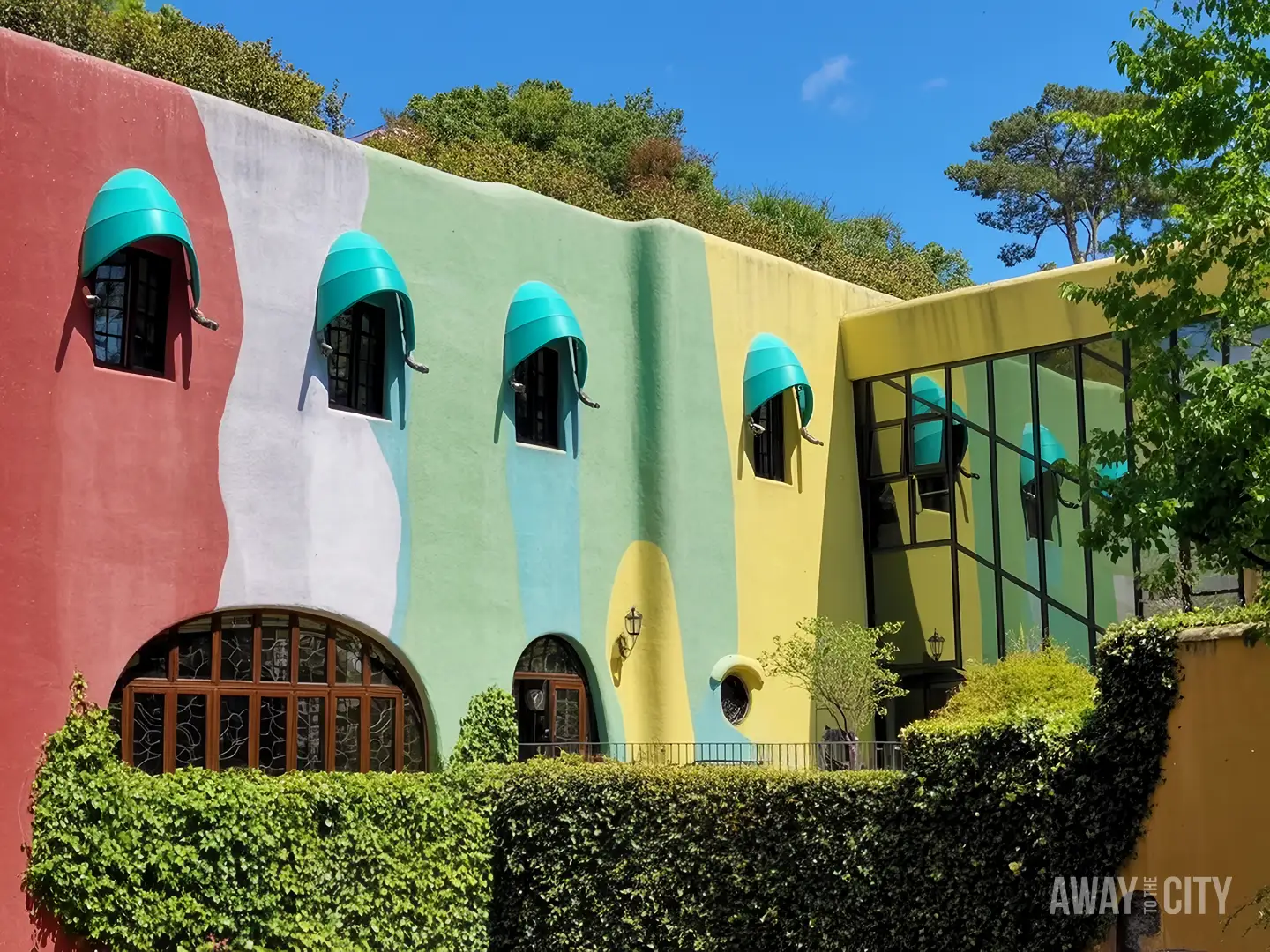 Colourful whimsical building with wavy pastel walls and teal window awnings, surrounded by greenery under clear blue sky.