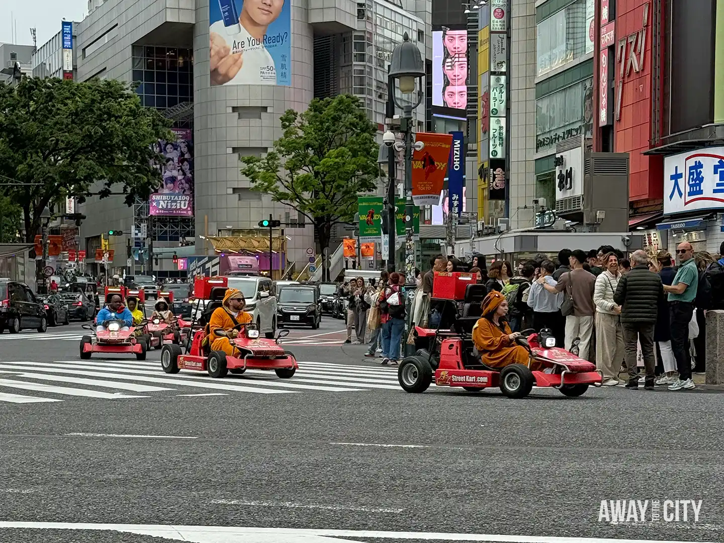 Tourists driving small red go-karts through busy Shibuya intersection while crowds watch from pavement, surrounded by tall buildings and billboards.