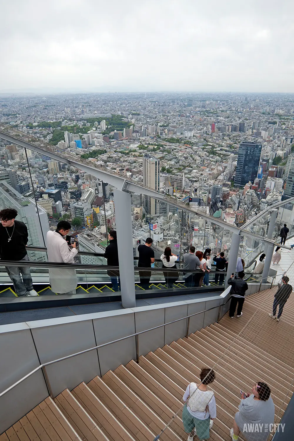 Visitors standing on wide wooden steps and glass-sided escalator at rooftop observation deck overlooking sprawling Tokyo cityscape under cloudy sky.