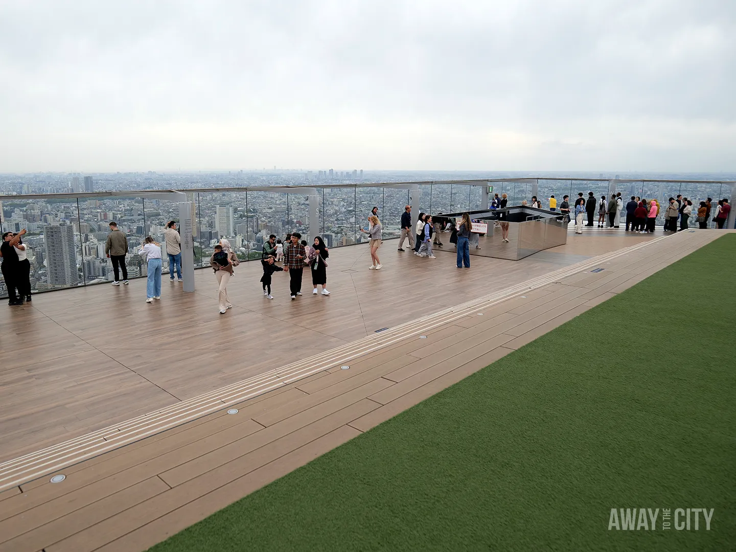 People walking and taking photos on spacious rooftop terrace with glass railing and artificial grass, overlooking vast Tokyo skyline.