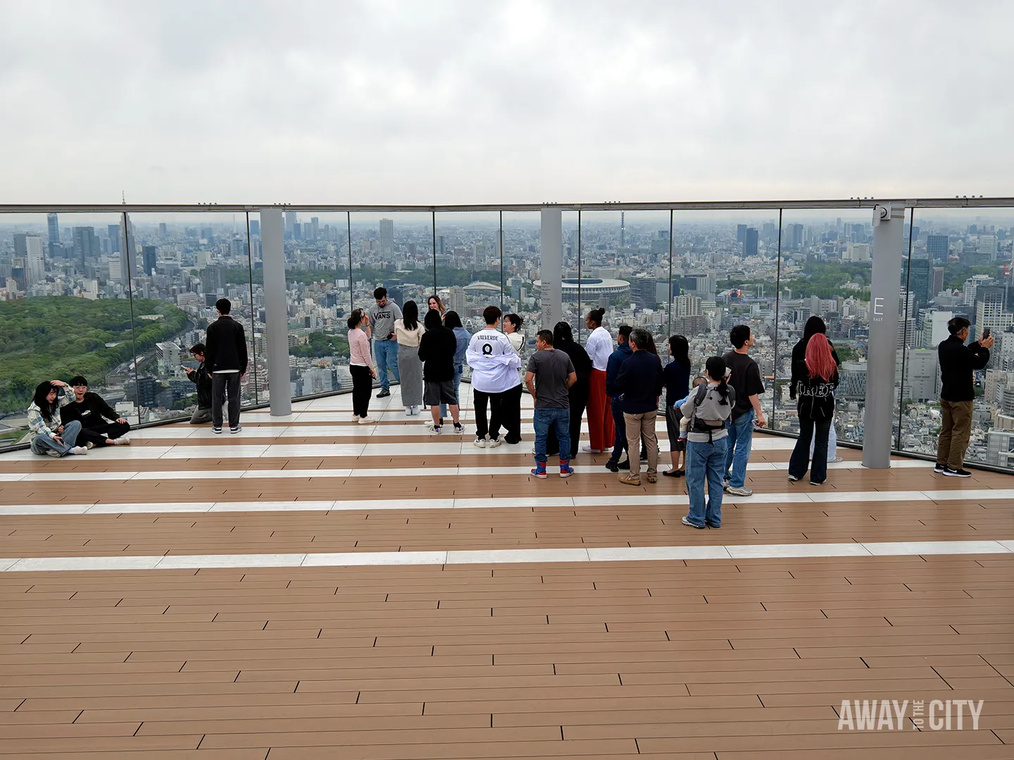 Crowd gathered along glass barrier at rooftop observation deck looking across Tokyo skyline, with Yoyogi Park greenery and stadium visible.