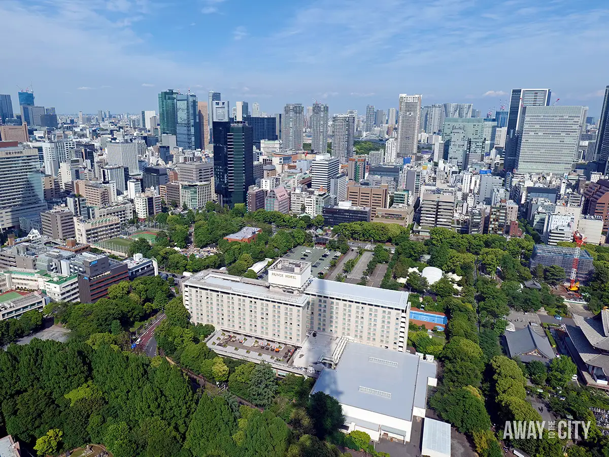 Aerial view of sprawling city skyline surrounding large green park and building labelled Prince Hotel, with dense towers beyond.