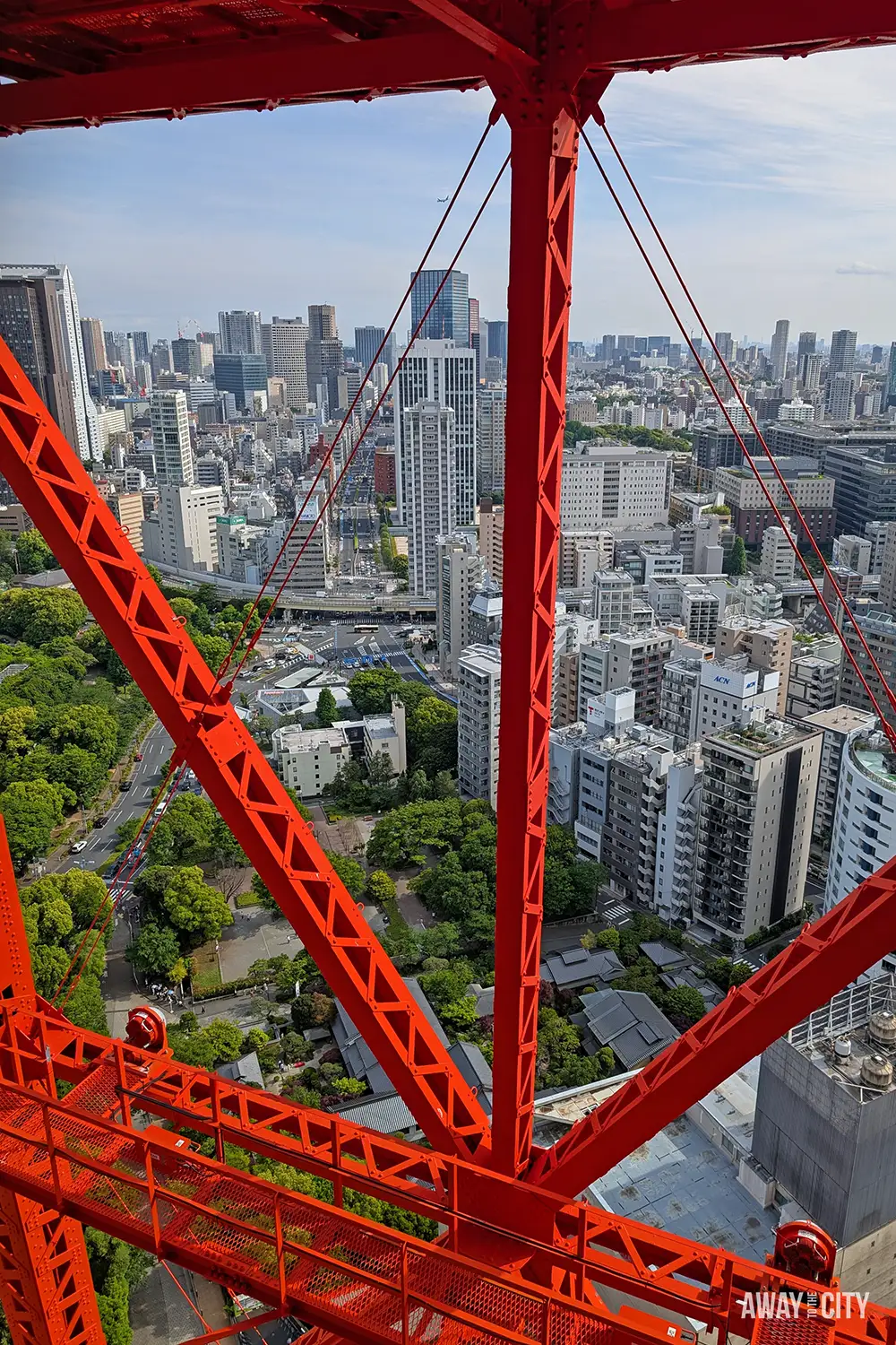 View through bright red steel lattice of observation tower framing busy city streets, parks, and high-rise buildings stretching to horizon.