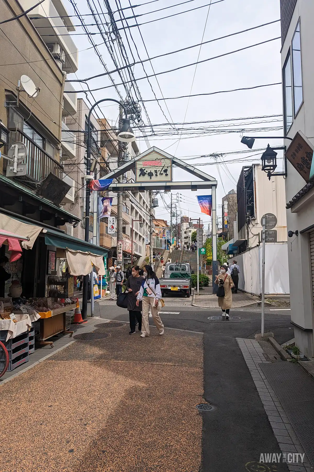 Entrance arch to Yanaka Ginza shopping street with Japanese text sign, overhead power lines, small stalls, and pedestrians walking.