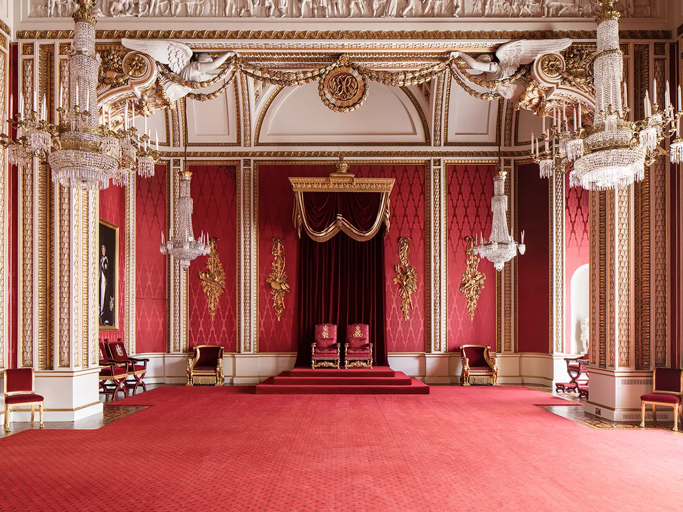 The richly decorated throne room at Buckingham Palace, with a long red carpet leading to two ornate red chairs.
