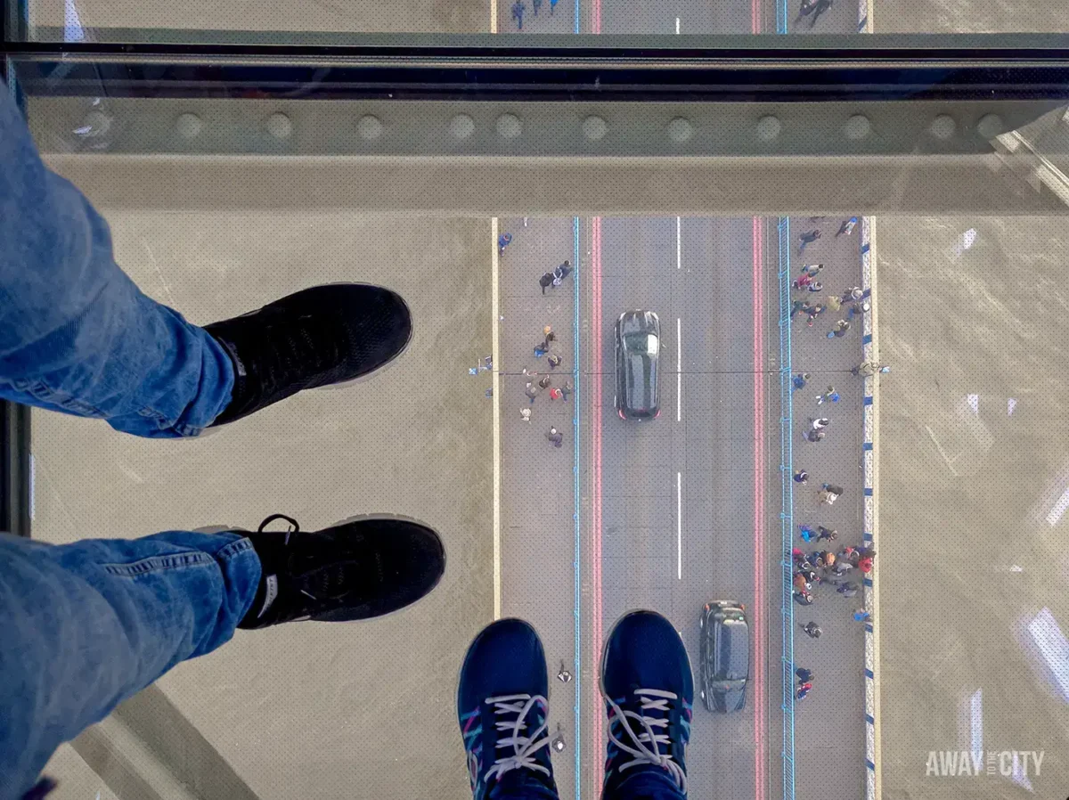 A pair of legs in jeans and trainers stands on the glass floor of Tower Bridge, looking down at the road and pedestrians below.