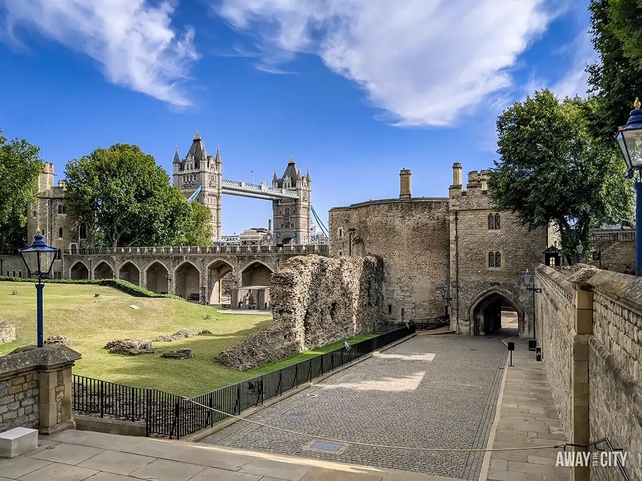 The inner courtyard of the Tower of London, with cobblestone paths, ancient stone walls, and Tower Bridge in the background.