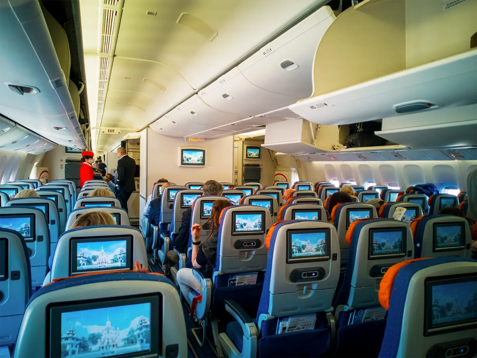 Interior of a passenger aeroplane cabin showing rows of economy seats, overhead lockers and seatback entertainment screens.