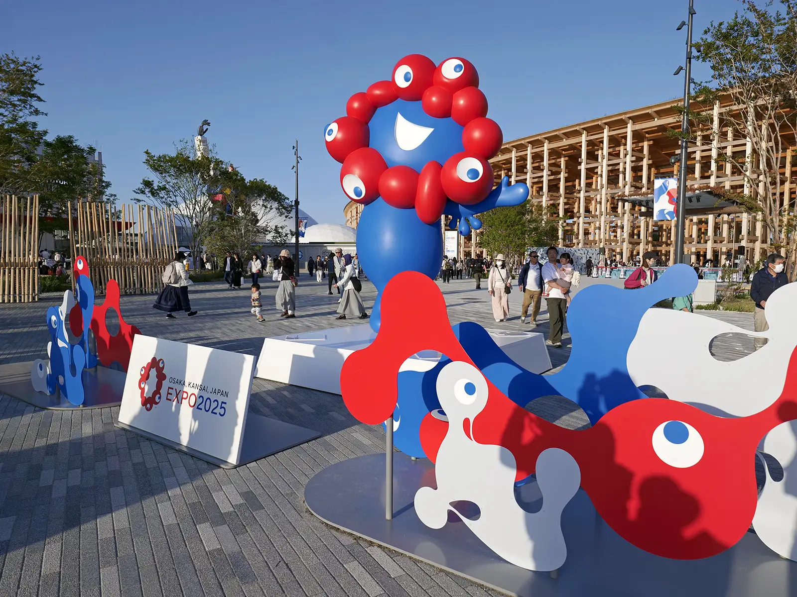 Myaku-Myaku, the blue and red character, is seen with other colourful shapes and Osaka Kansai Japan Expo 2025 signage outdoors.