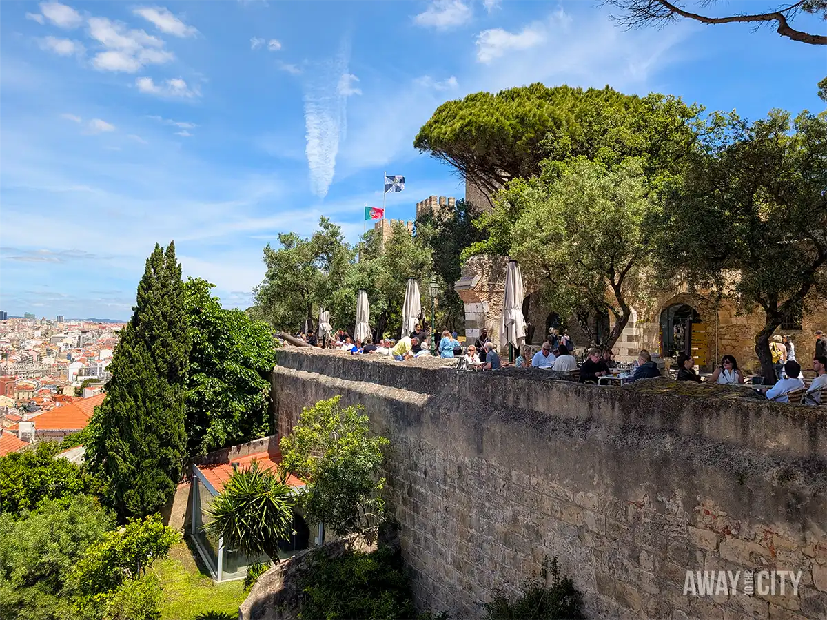 Visitors relax on a terrace at Castelo de São Jorge (Saint George’s Castle) in Lisbon, beside historic stone walls and trees.