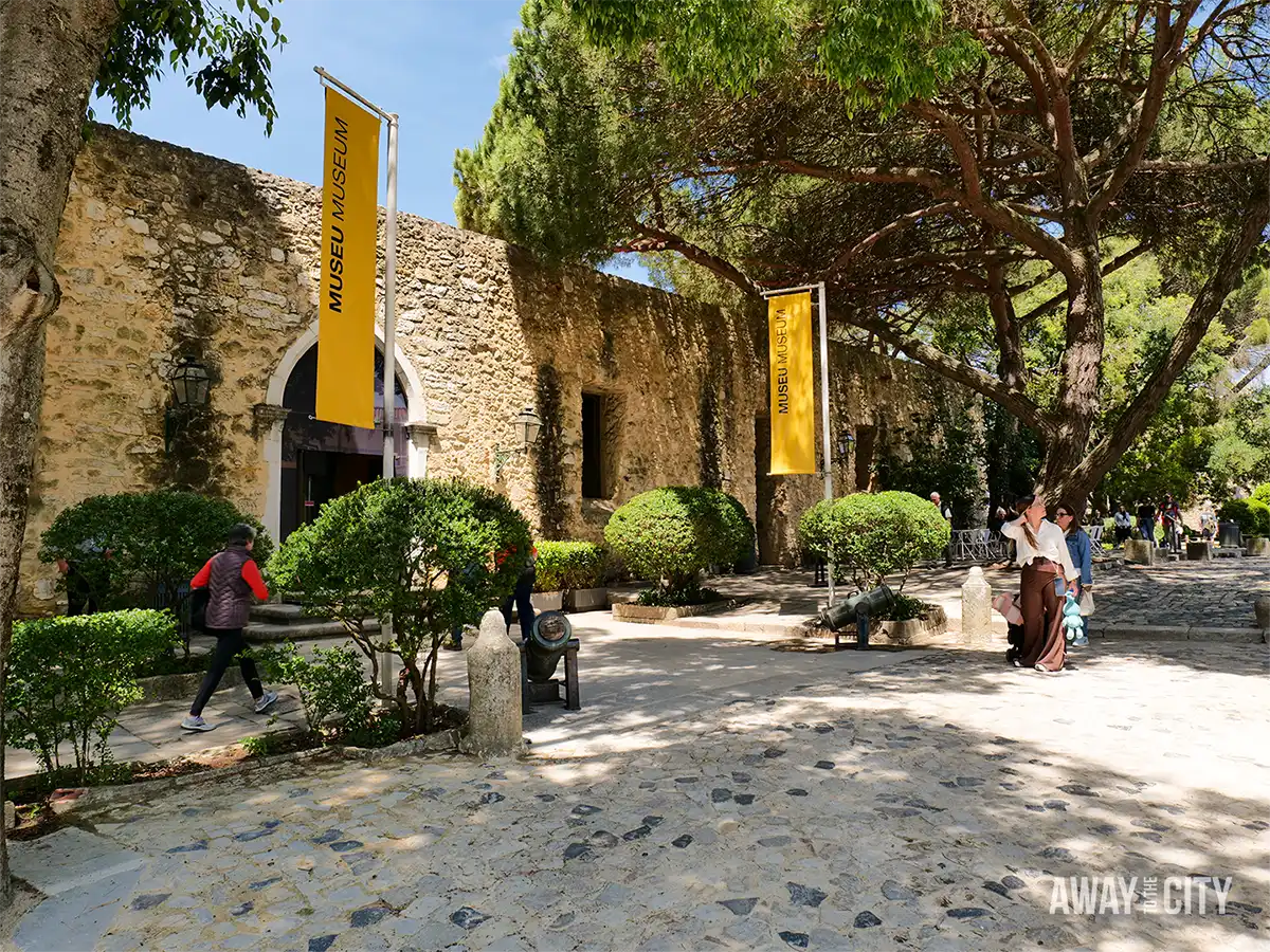Remains of the original Royal Palace of the Alcáçova and shaded courtyard at Castelo de São Jorge (Saint George's Castle) in Lisbon, with visitors strolling among trees.