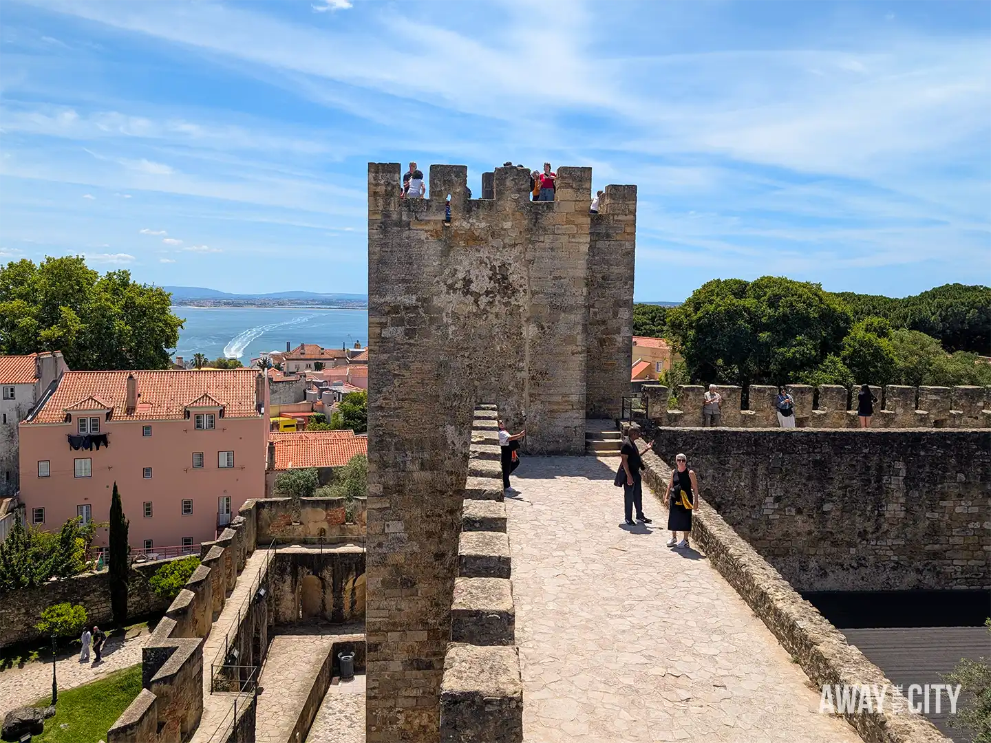 Visitors walk along medieval battlements and tower at Castelo de São Jorge (Saint George's Castle) in Lisbon, overlooking the city skyline.