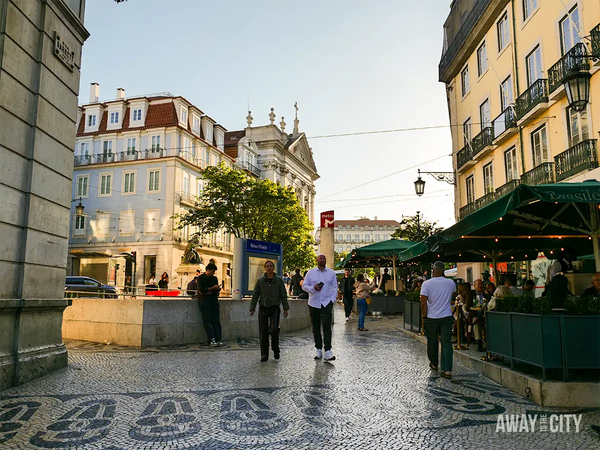 People walk across a cobbled square lined with cafés and historic buildings in Chiado, Lisbon, during late afternoon.