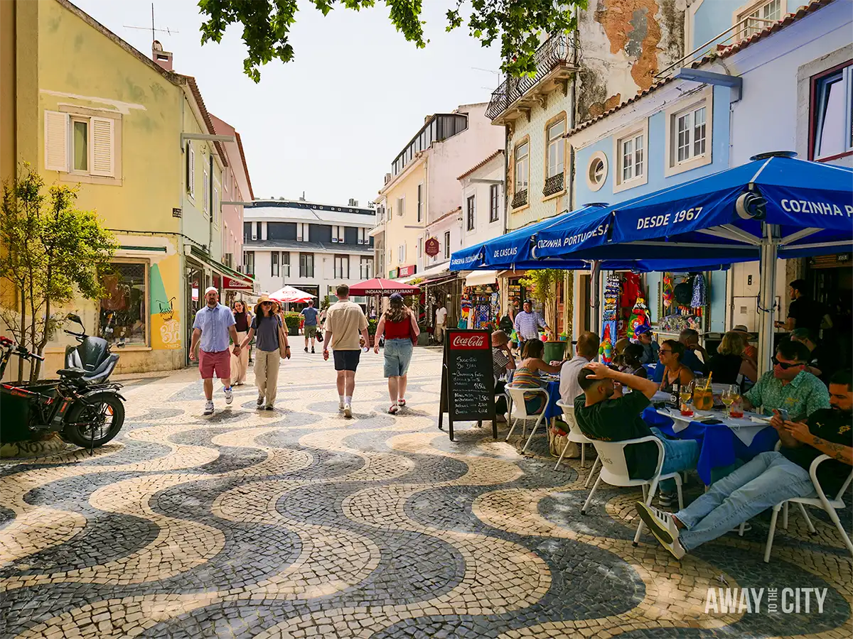 Pedestrians stroll along Rua Frederico Arouca in Cascais, lined with cafés, colourful buildings and patterned pavements.