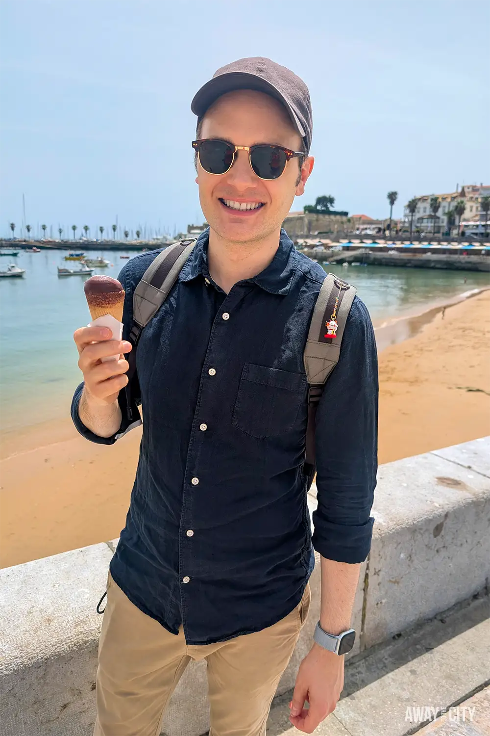 Person enjoying an ice cream beside Praia da Ribeira de Cascais beach, with boats and the waterfront in the background.