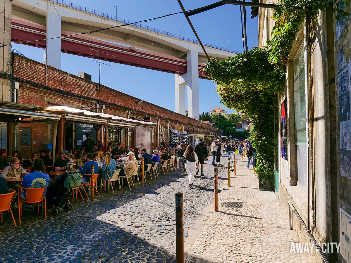 Outdoor bars and visitors line a cobbled street at LX Factory in Lisbon, beneath industrial buildings and elevated structures.