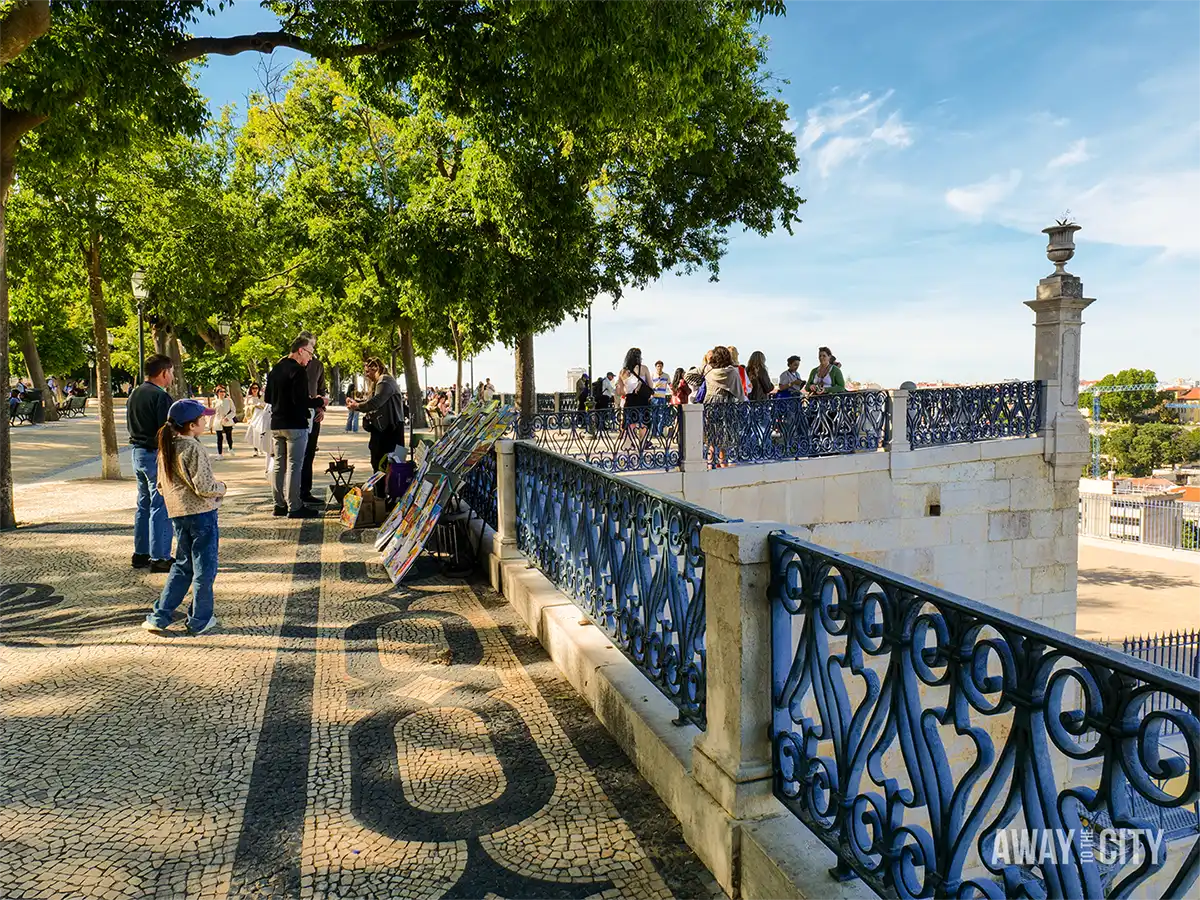 Visitors gather on the viewing platform at Miradouro de São Pedro de Alcântara in Lisbon, beneath leafy trees and ornate railings.