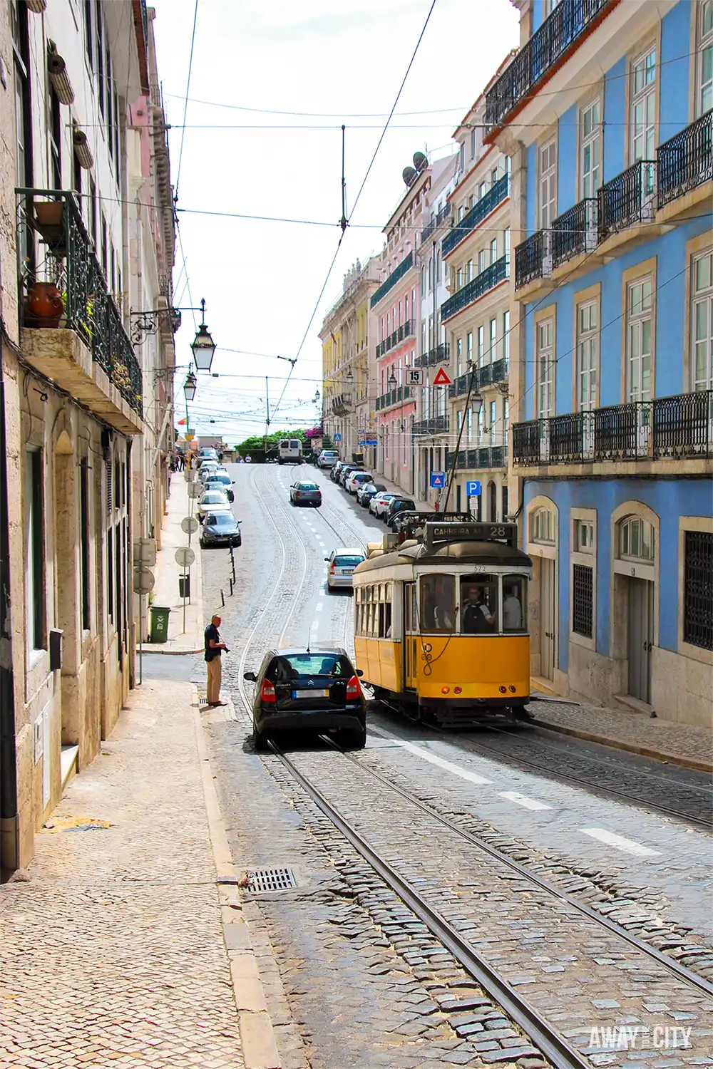 Historic yellow Tram 28 travels uphill along a cobbled street lined with pastel buildings in Lisbon.
