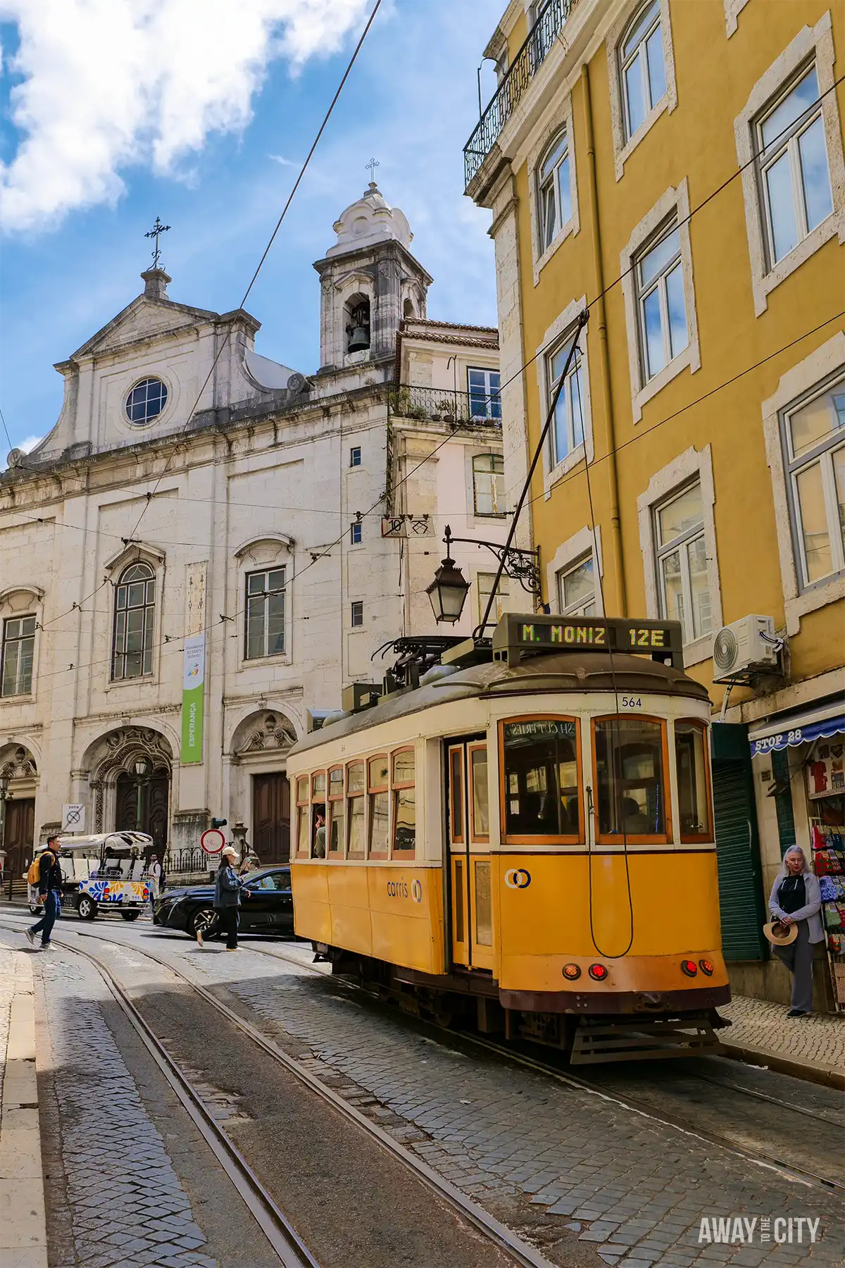 Historic yellow Lisbon tram travels past a church along a narrow street lined with traditional buildings.