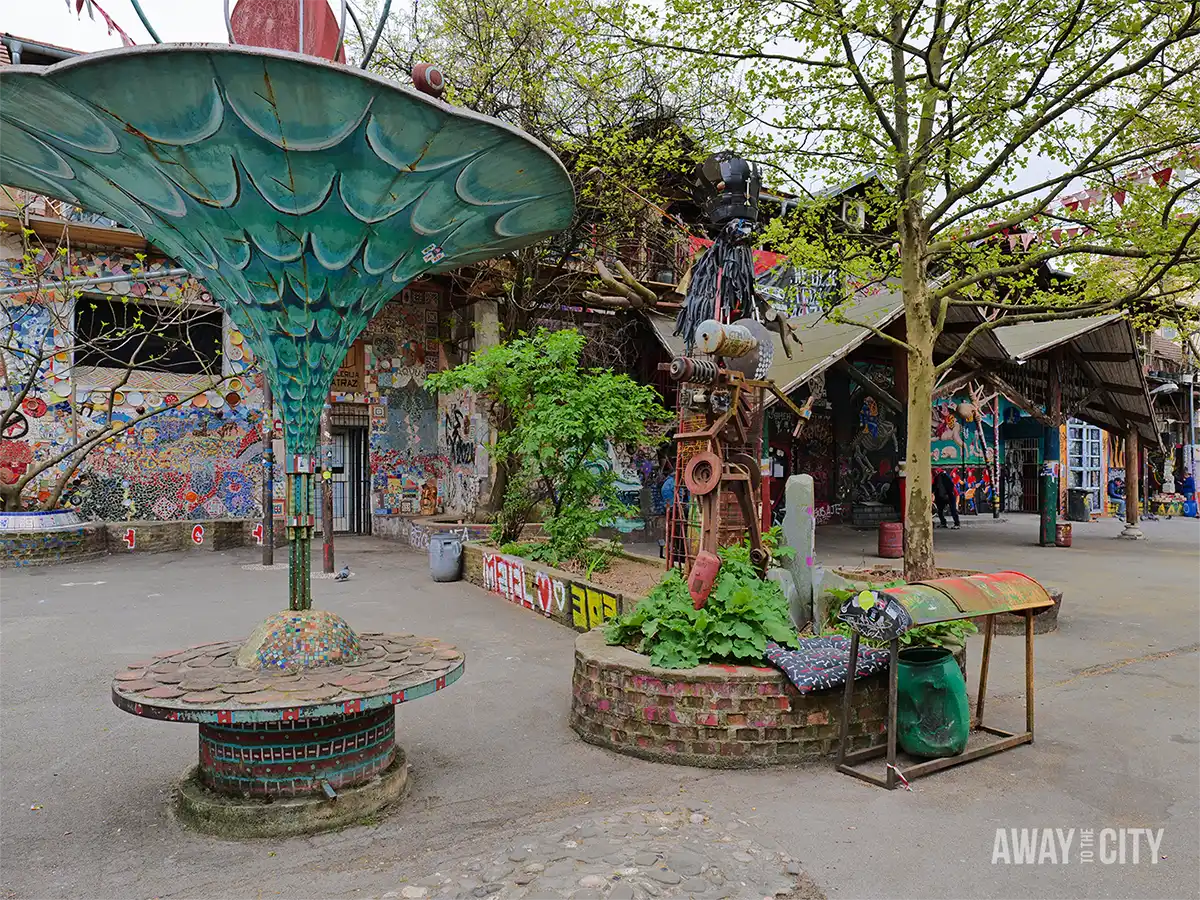Open courtyard at AKC Metelkova Mesto featuring quirky sculptures, mosaic structures, trees, and vibrant street art across surrounding alternative cultural buildings.