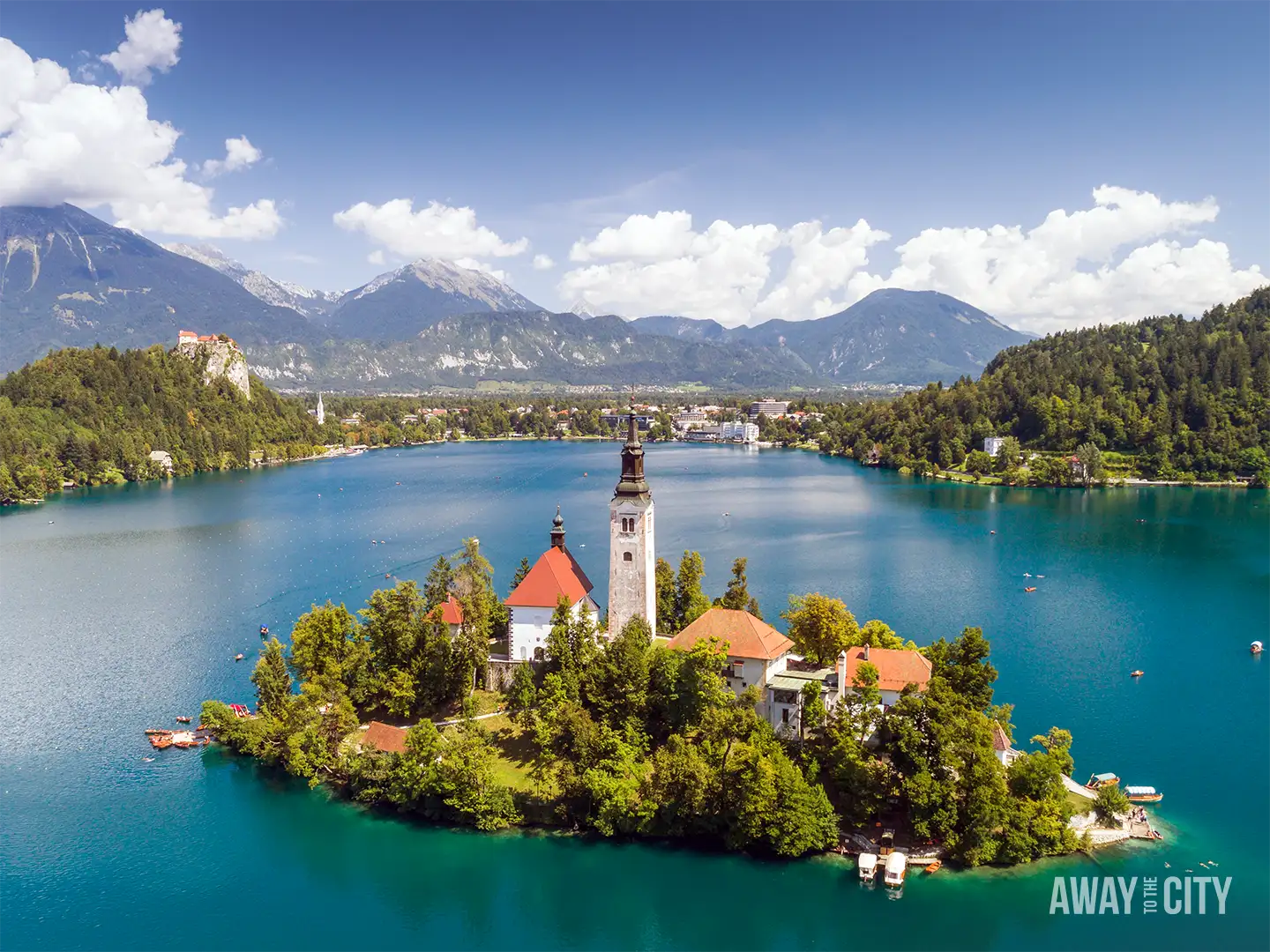Aerial view of Lake Bled with island church and bell tower, surrounded by turquoise water, forested hills, and distant Alpine mountains under blue sky.