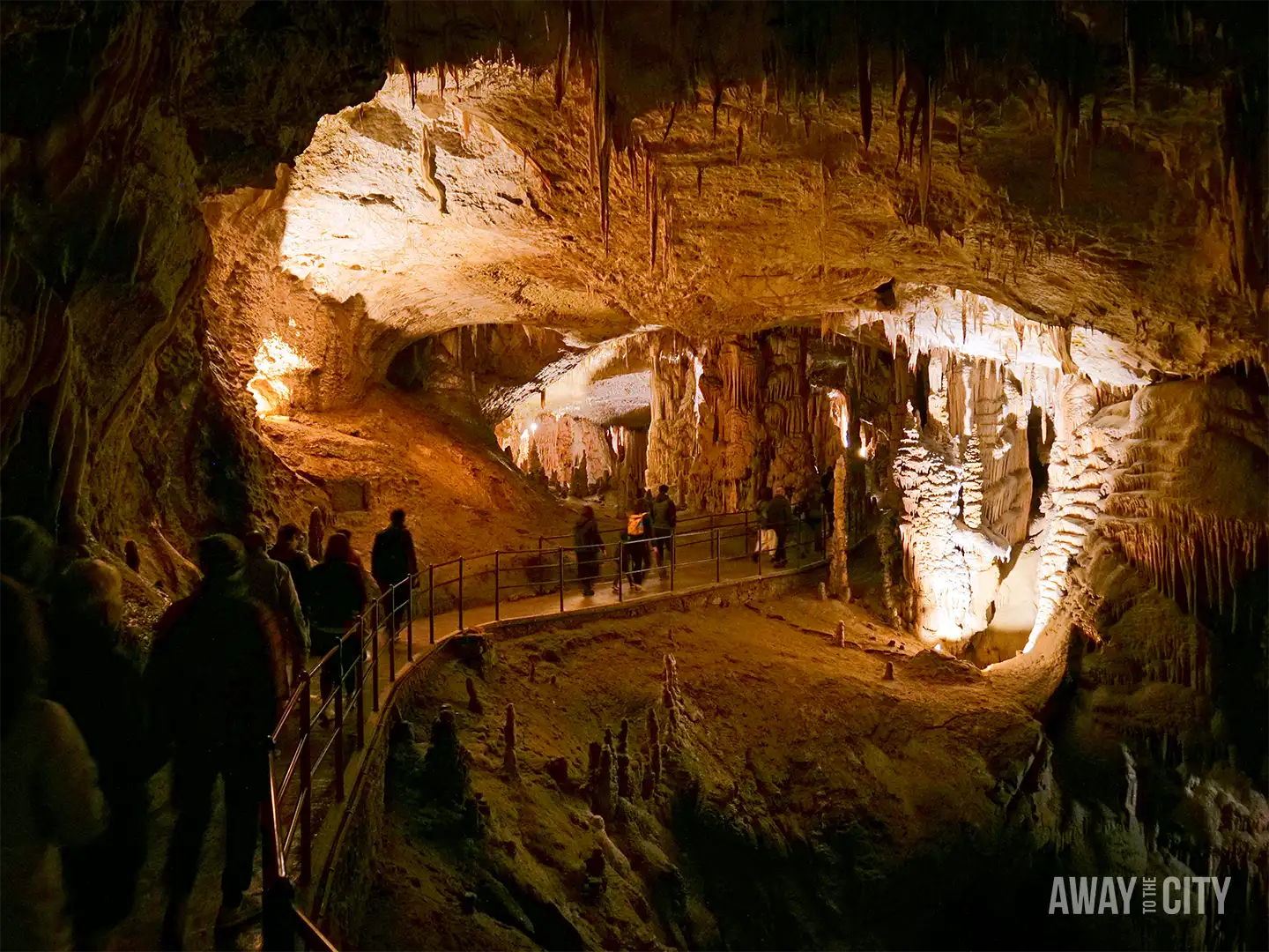 Visitor standing inside Postojna Cave beside towering rock formations, with crowds exploring vast chamber filled with stalactites and textured limestone walls.
