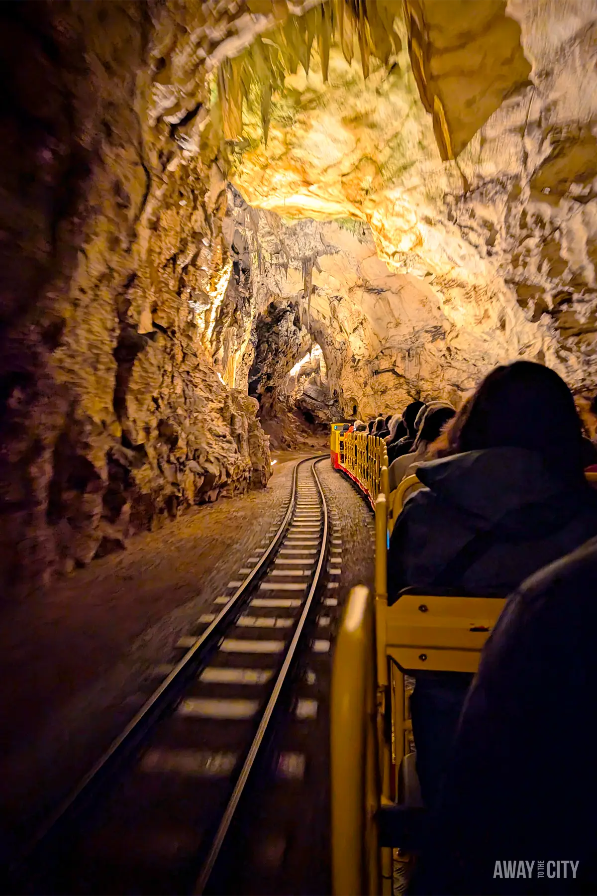 Tourists riding small train through Postojna Cave tunnel, surrounded by rocky formations and illuminated cavern walls along narrow railway track.