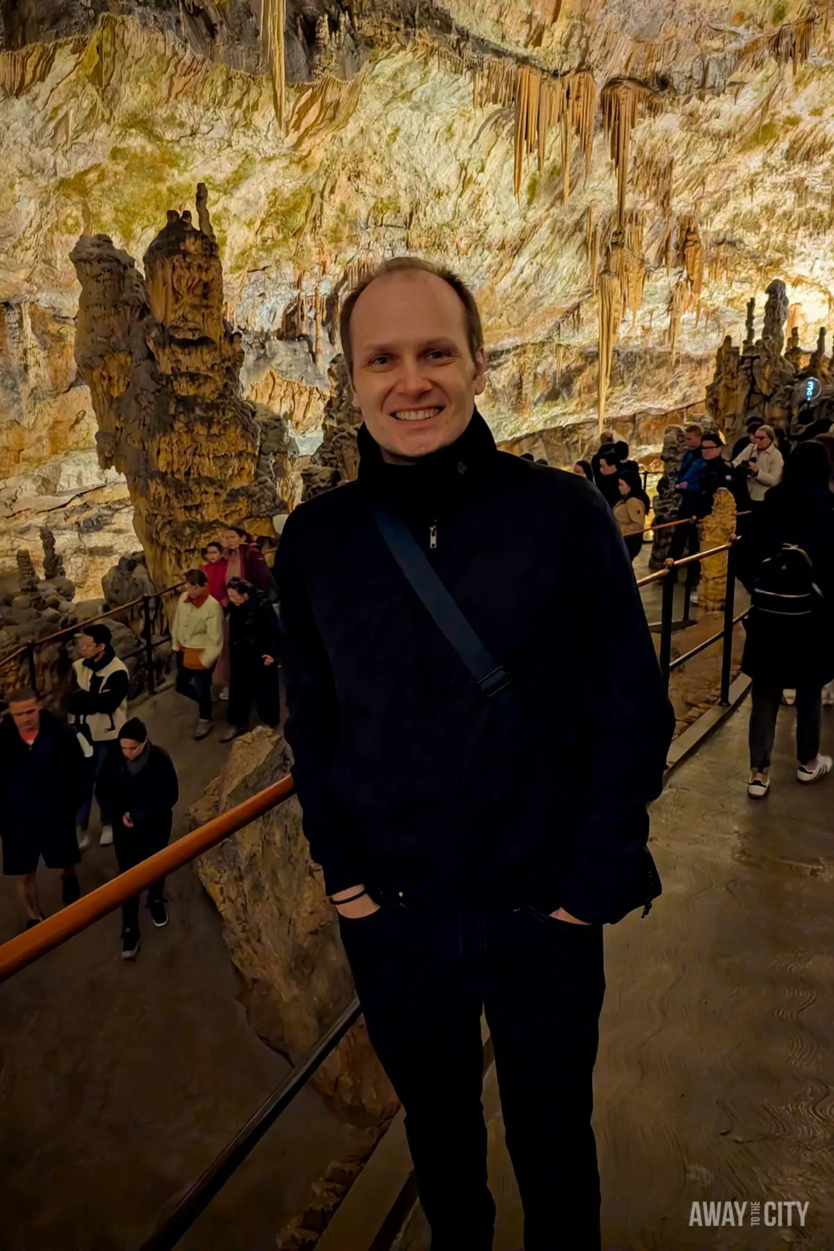 Interior of Postojna Cave with visitor walkway, dramatic stalactites and stalagmites, and warm lighting illuminating vast underground chambers during guided tour.