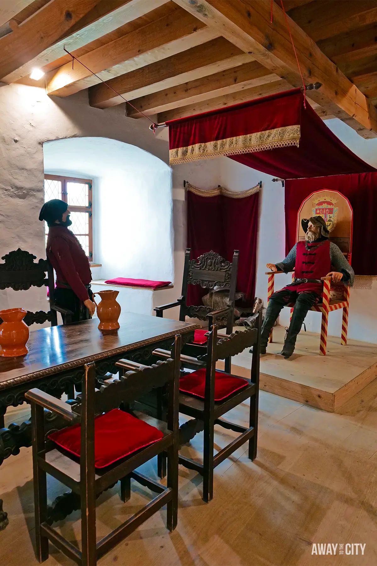 Interior of Predjama Castle with medieval-style room, wooden beams, red canopy bed, and mannequins dressed in period clothing.