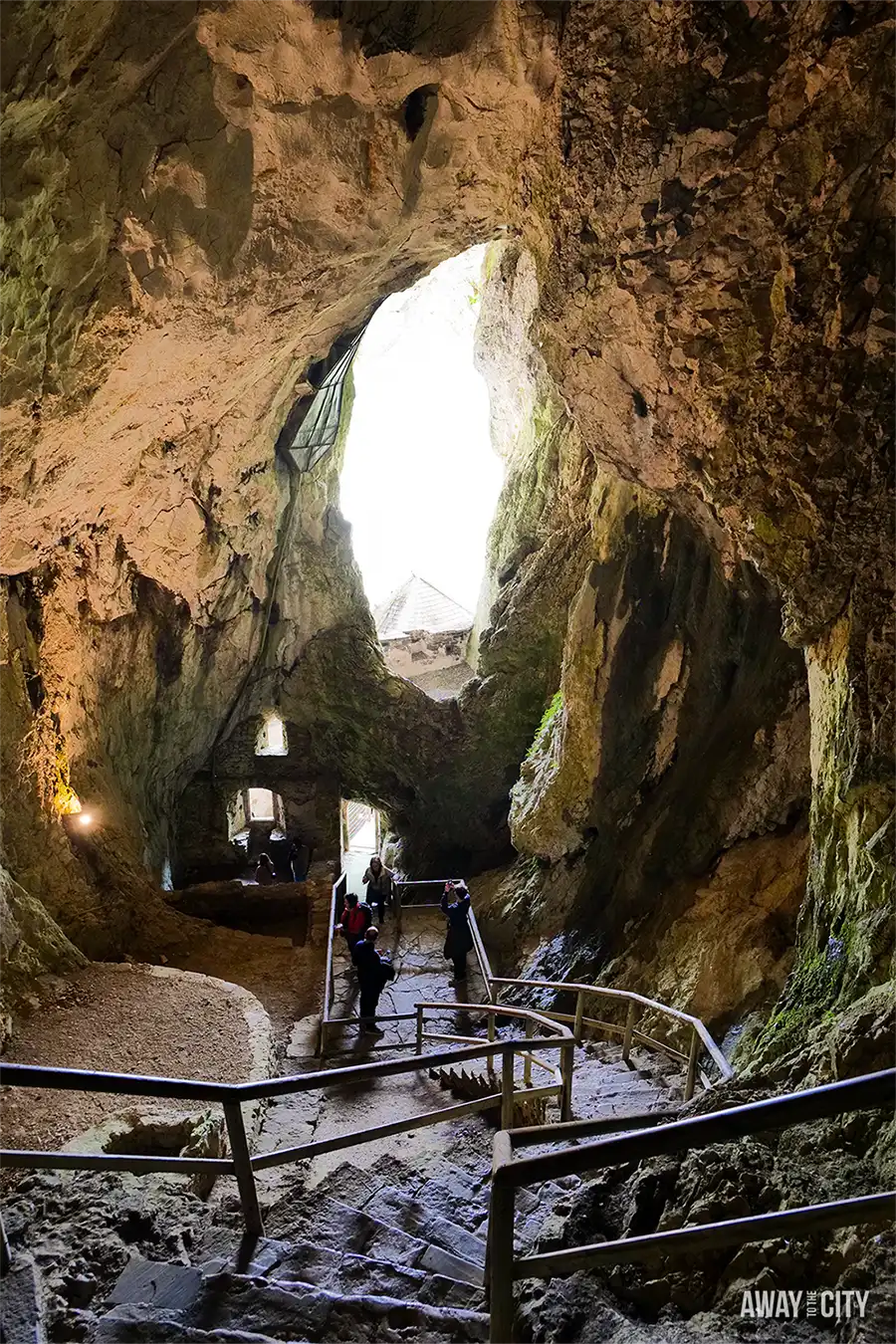 Large cave chamber behind Predjama Castle with rocky walls, natural light from opening, and visitors walking along stepped pathway.