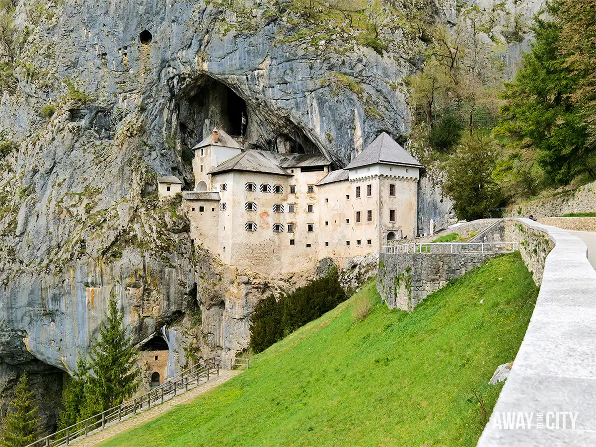 Predjama Castle built into towering cliff face with cave opening above, surrounded by green hillside and stone pathway leading towards entrance.