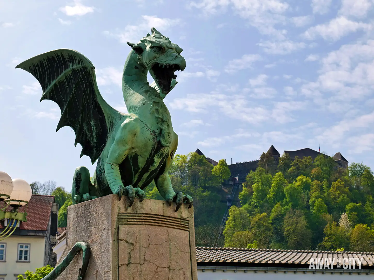 Close view of Ljubljana Dragon Bridge statue with green winged dragon perched on stone pedestal, castle hill rising in background under blue sky.