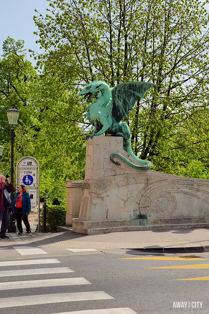 Wide street view of Dragon Bridge showing dragon statue, pedestrians crossing road, and urban surroundings on a bright day, one of the best things to see in Ljubljana, Slovenia.