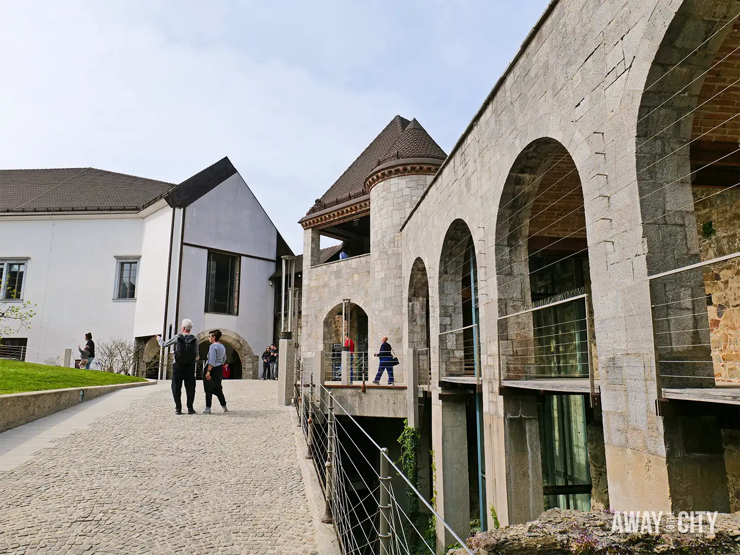 Ljubljana Castle courtyard with stone arches and modern walkway, visitors strolling along cobbled path beside historic fortress walls.
