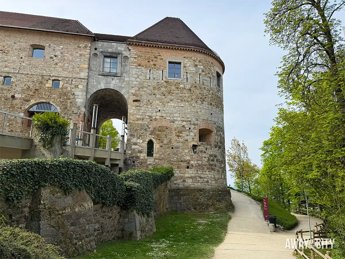 Ljubljana Castle exterior with round stone tower, arched entrance, and pathway leading uphill, surrounded by greenery under overcast sky.