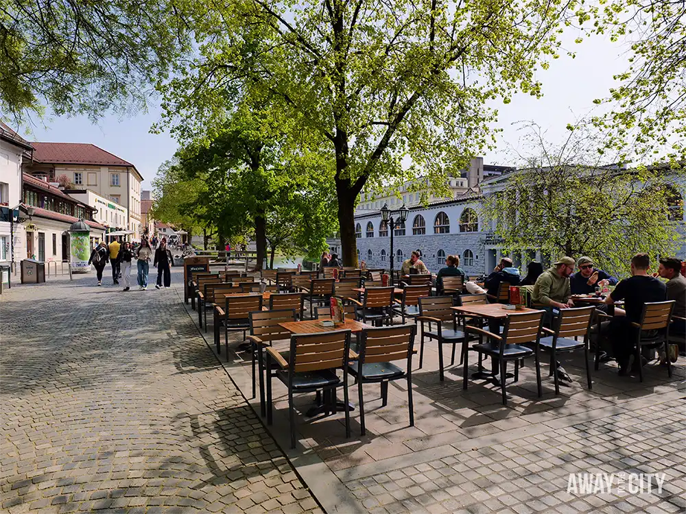 Outdoor café seating along Ljubljanica promenade with wooden tables under leafy trees, people dining, and cobbled walkway beside the river.