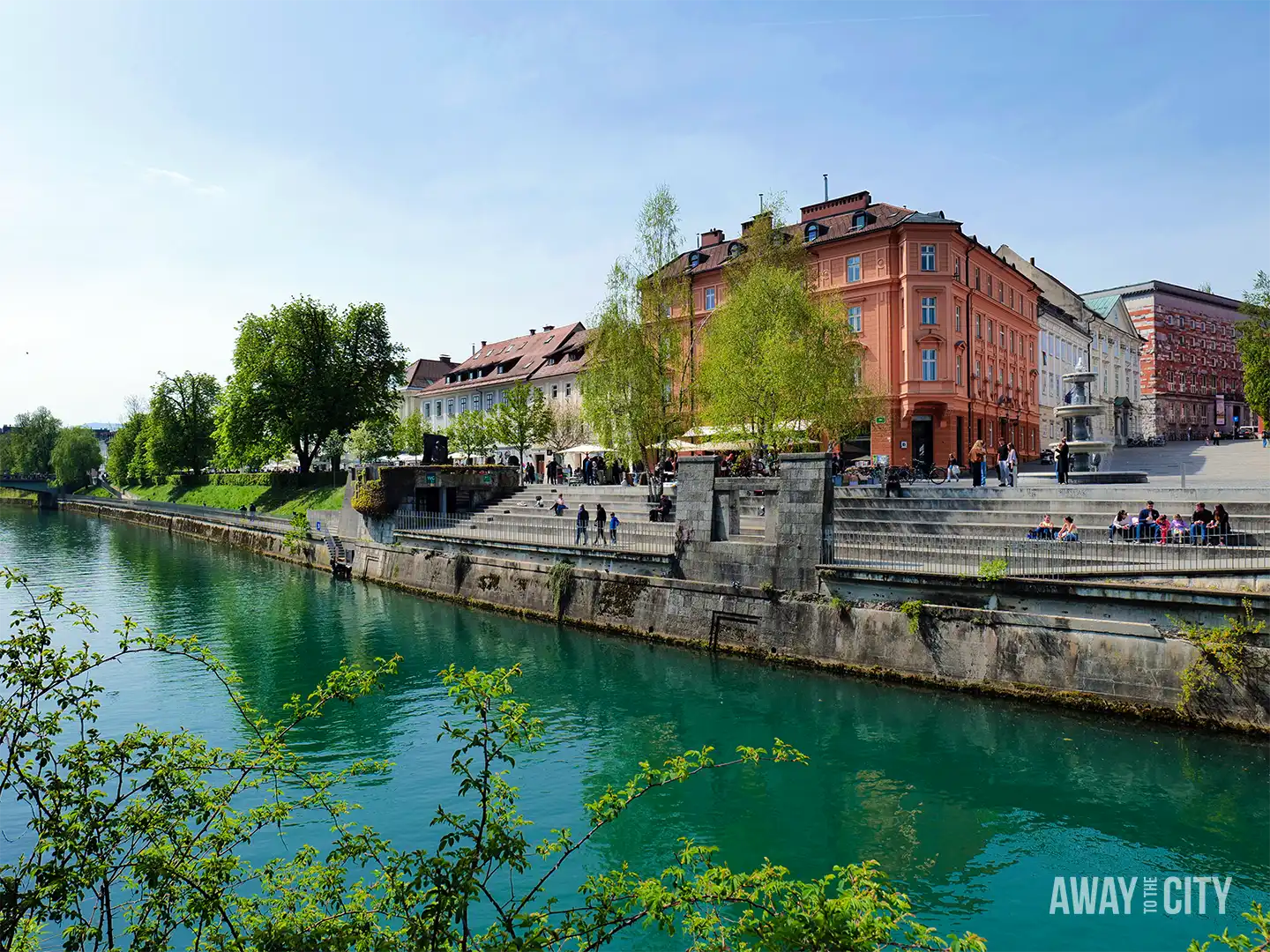 Riverside view along Ljubljanica promenade with green water, pastel buildings, trees, and people strolling beside cafés on a sunny day.