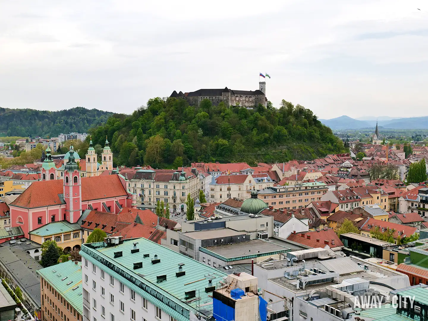 View from Nebotičnik Skyscraper over Ljubljana with red rooftops, Franciscan Church, and castle on hill, with construction crane in foreground.