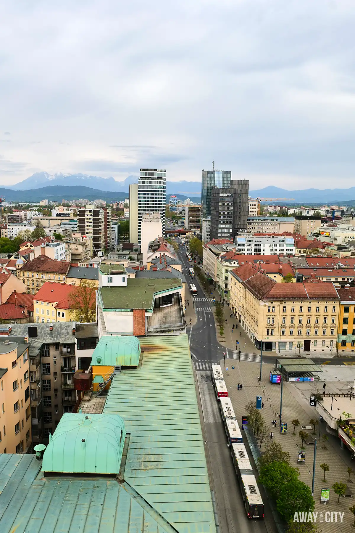 Elevated city view from Nebotičnik Skyscraper showing Ljubljana rooftops, streets with tram, and distant hills under overcast sky.
