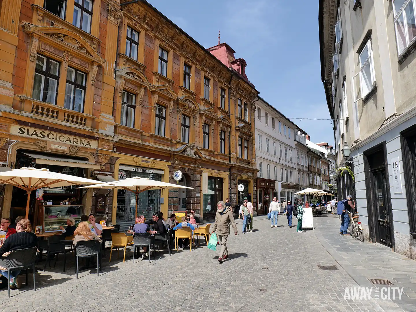 Gornji Trg street in Ljubljana lined with historic buildings, outdoor café tables, pedestrians walking, and ornate facades, one of the must-see spots in Slovenia's capital.