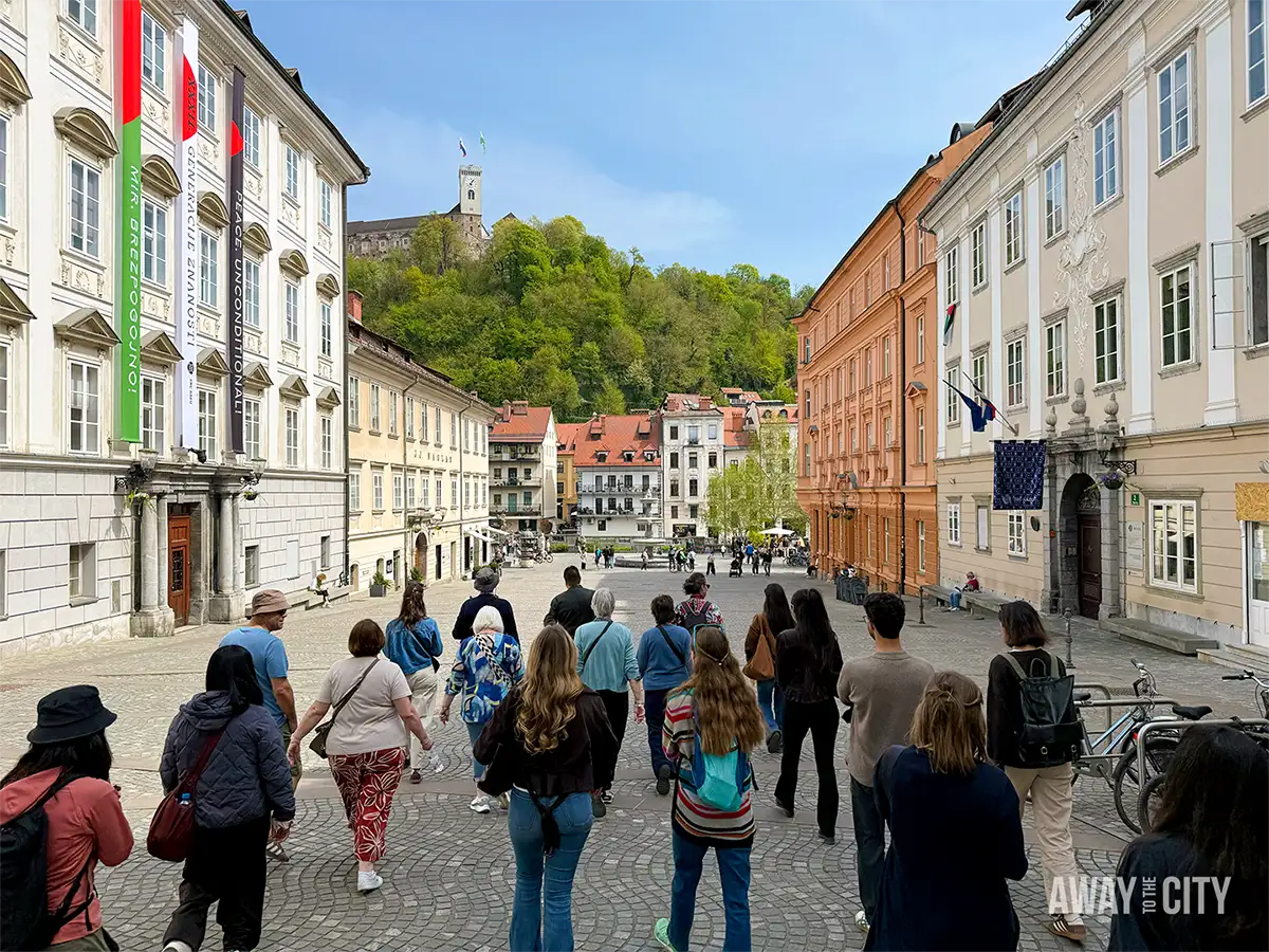 Walking tour group in Ljubljana strolling through cobbled Old Town square, flanked by historic pastel buildings with castle hill rising in the background.