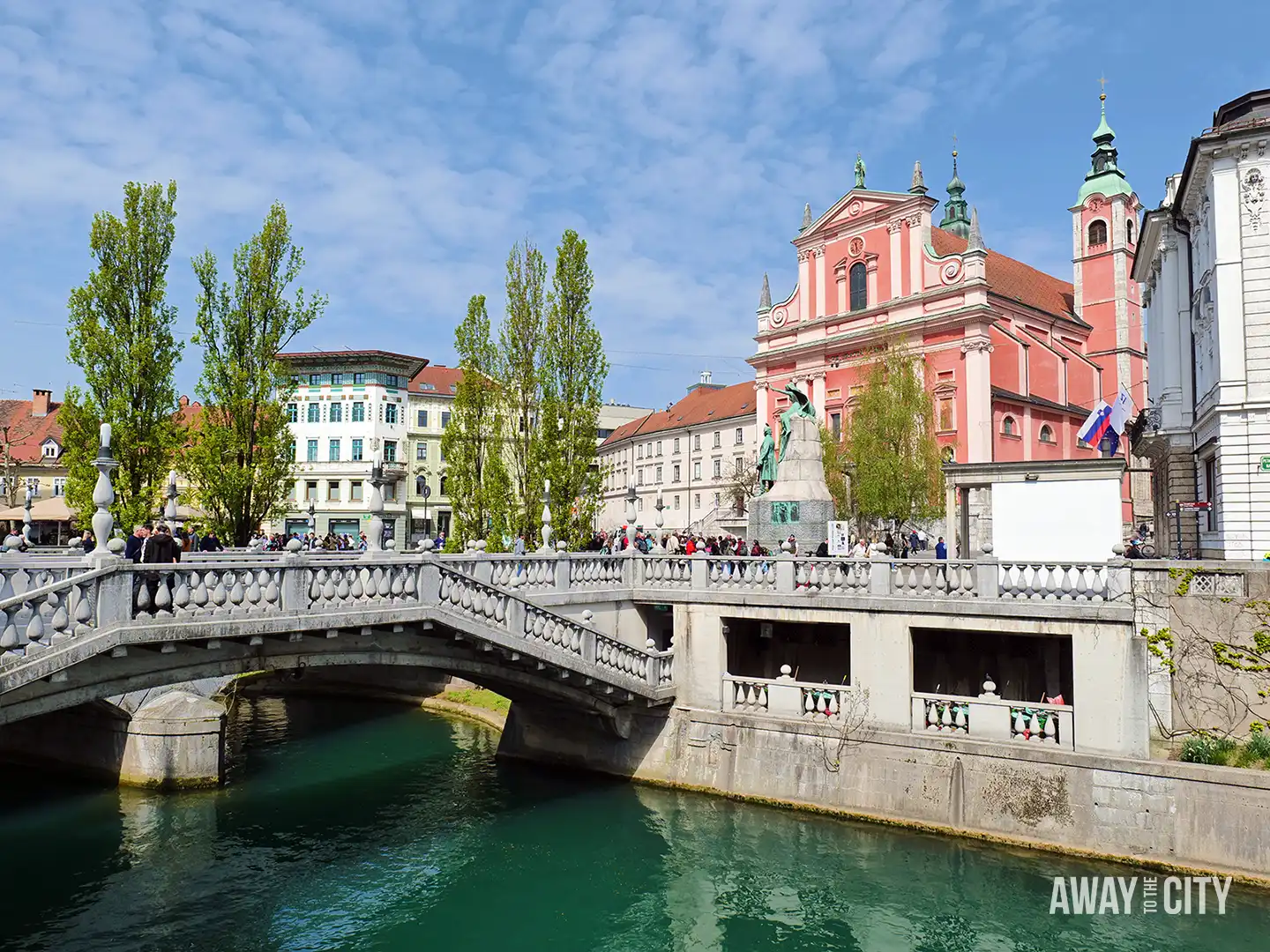 Prešeren Square in Ljubljana with stone bridge over green river, pink Franciscan church, statue monument, and crowds gathered along the riverside promenade. It's one of the best things to do in Ljubljana.