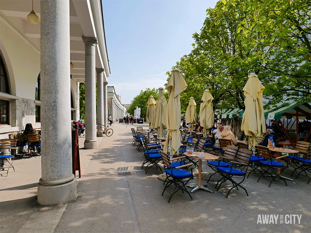 Riverside café terrace on Adamič-Lunder embankment with rows of chairs under folded umbrellas beside a colonnade.